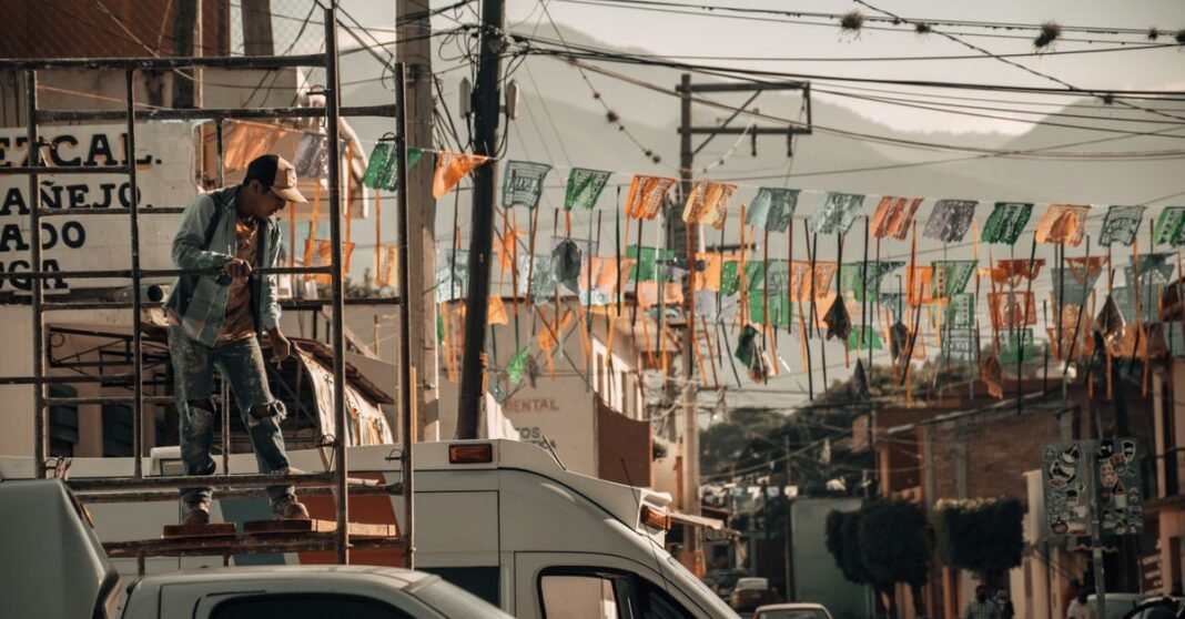 Kia invertirá 649 millones de dólares en Nuevo León para fabricar el EV3 eléctrico Trabajador de la construcción en un andamio en una vibrante calle de la ciudad, rodeado de banderas coloridas.
