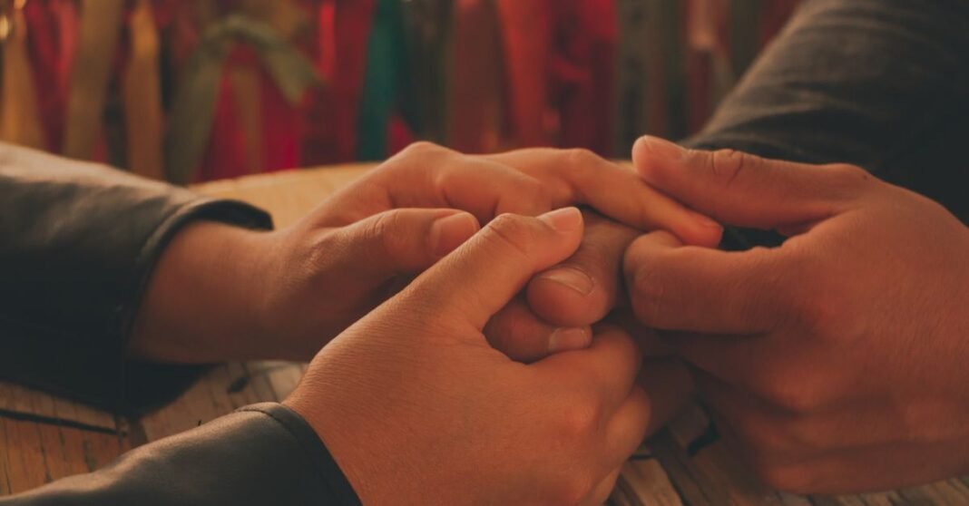 UdeG y notarios de Jalisco formalizan programa para donar cuerpos con fines de docencia e investigación Acercamiento cálido e íntimo de una pareja cogiendo las manos sobre una mesa de madera con cintas de colores de fondo.