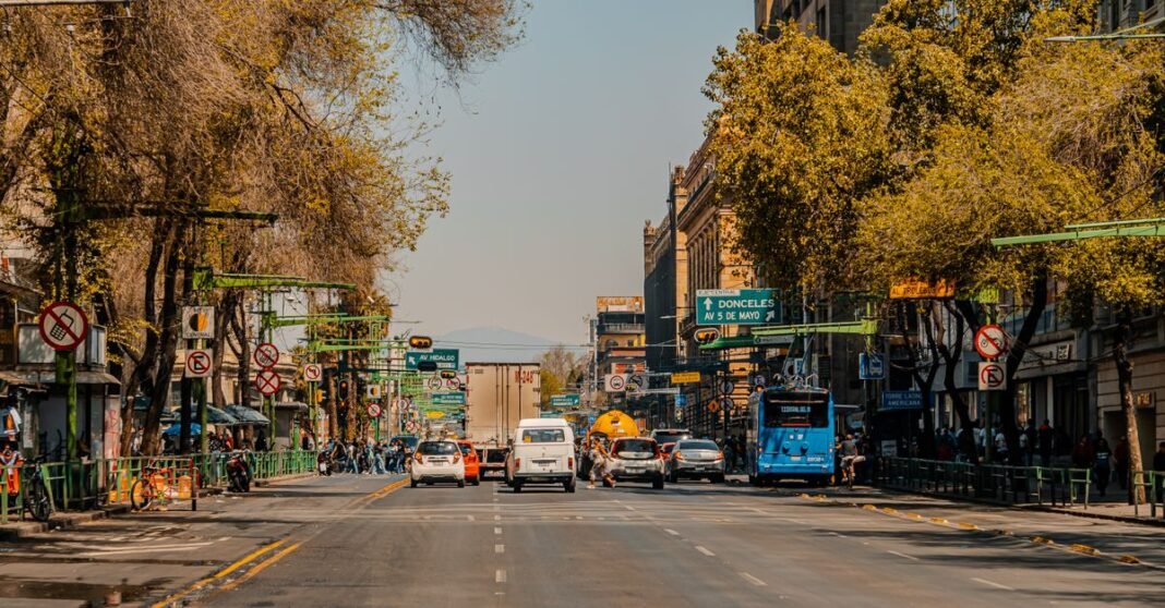 Columna plantea medir el polvo urbano en CDMX; cifras citadas requieren precisar su año de referencia Coches y peatones en una calle urbana concurrida en Ciudad de México, bordeada de árboles y edificios.