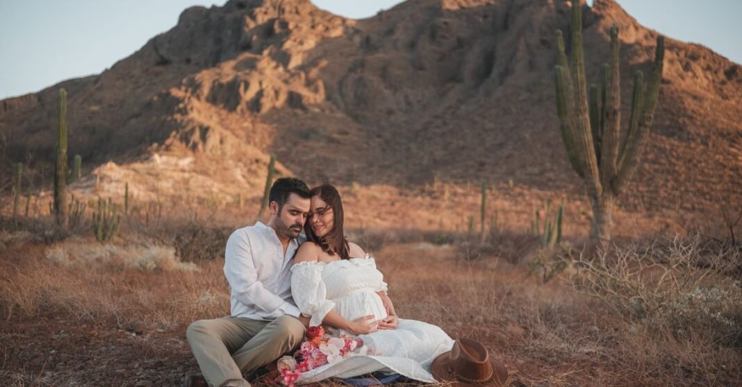 OMS impulsa la inclusión de embarazadas y mujeres lactantes en ensayos clínicos Una pareja embarazada amorosa sentada junta en un sereno paisaje desértico, abrazando la belleza de la naturaleza.
