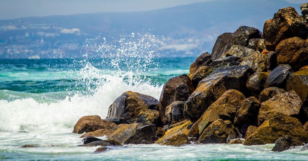 Tormenta tropical Iselle se forma lejos de México; prevén oleaje peligroso en Baja California Olas dinámicas del océano rompiendo contra rocas costeras en Ensenada, México, bajo un brillante cielo de verano.