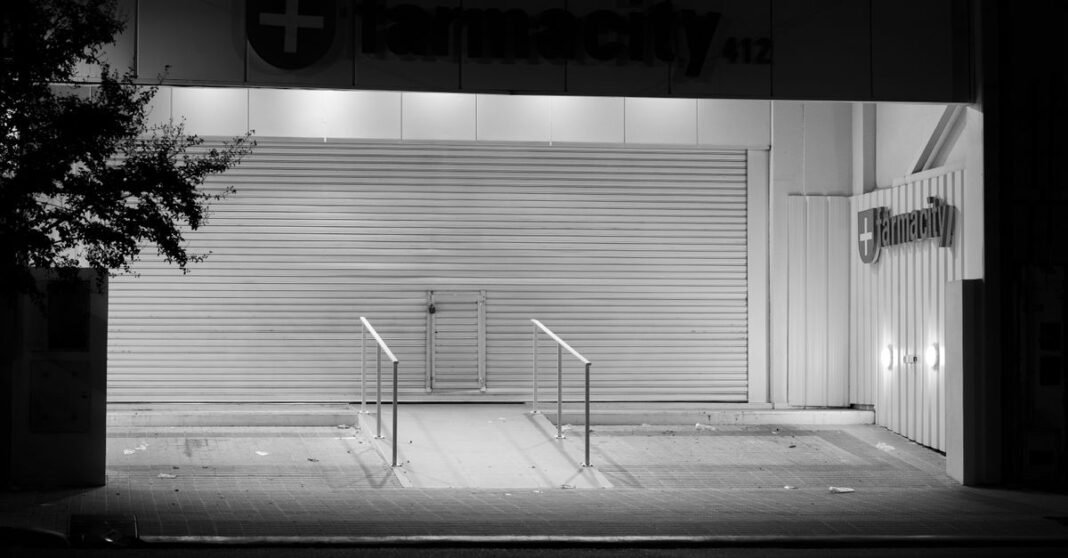 Persisten las críticas en Argentina a la reforma que limita a los médicos a prescribir por nombre genérico Fotografía en blanco y negro de una farmacia cerrada en Córdoba, Argentina, por la noche, en la que se aprecian persianas metálicas y un árbol.
