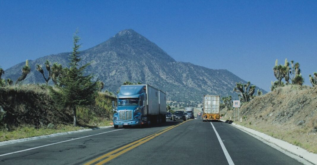 Trump aplaza un mes los aranceles a productos de México y parte de Canadá Camiones circulando por una carretera con un fondo de montaña bajo un cielo azul claro.