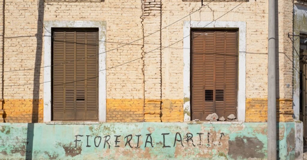 Fachada de ladrillo con persianas y base turquesa en Oliva, Córdoba, Argentina.