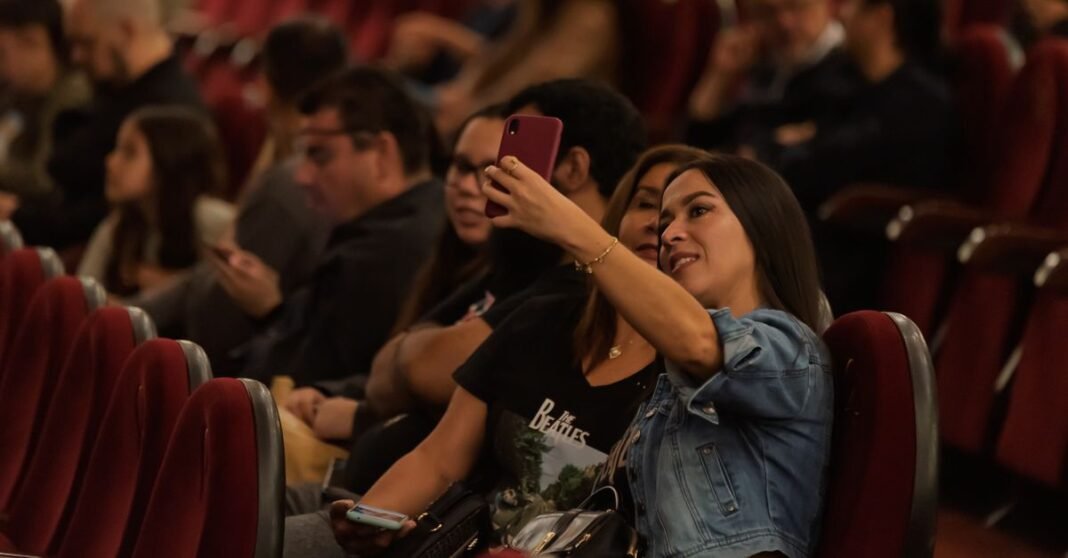 Un grupo de adultos en un teatro, se centra en dos mujeres que se toman una selfie en medio del público.
