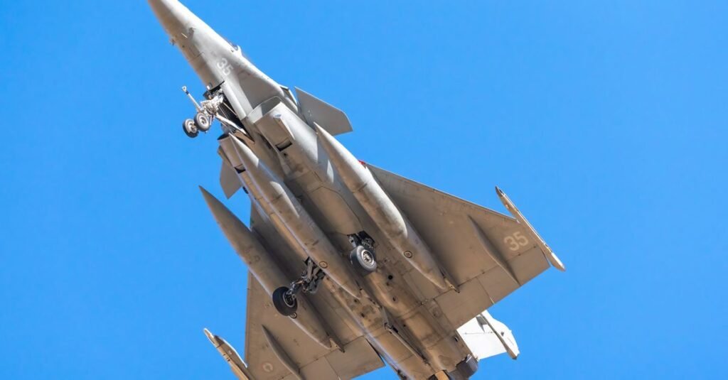 Un caza Rafale volando contra un cielo azul claro, visto desde abajo.
