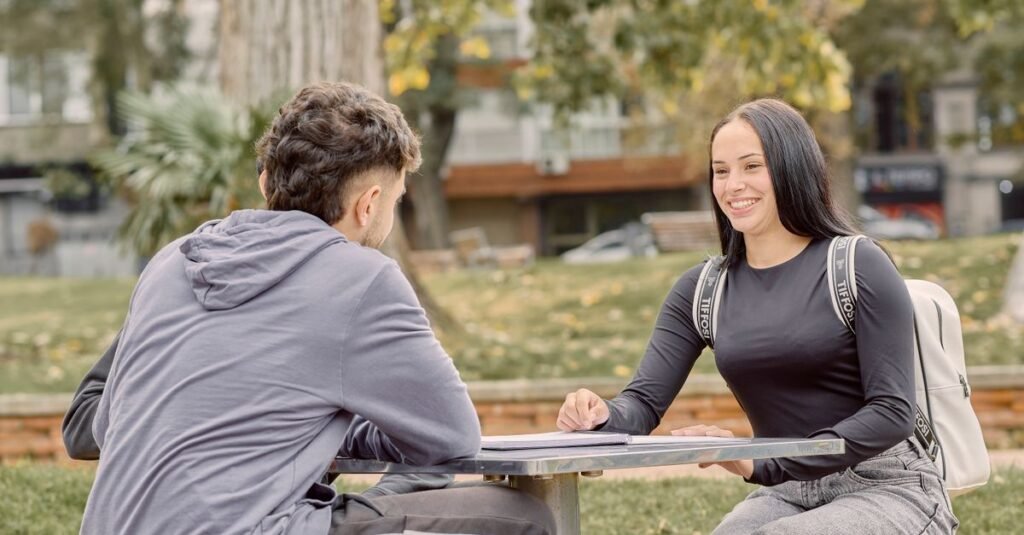 Dos jóvenes adultos sentados en una mesa al aire libre, sonriendo y conversando en un entorno de parque.