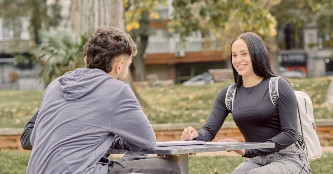 Dos jóvenes adultos sentados en una mesa al aire libre, sonriendo y conversando en un entorno de parque.