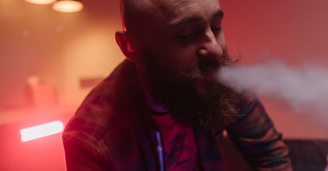 BAT México suspende la venta de vapeadores en el país tras la prohibición constitucional Primer plano de un hombre calvo con barba exhalando vapor en un espacio interior con poca luz.