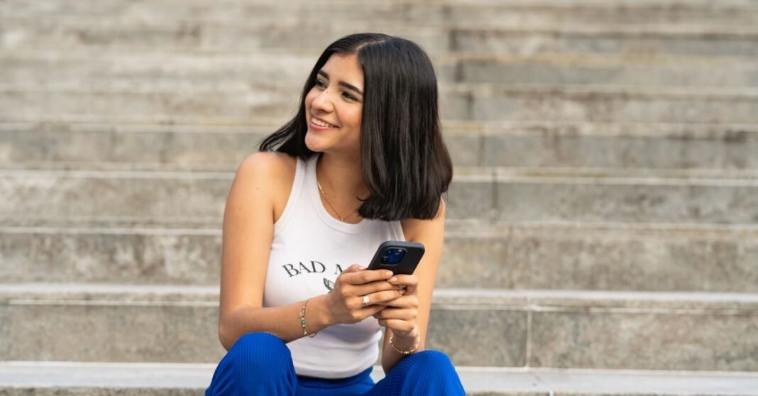 SEP anuncia restricción total de celulares en primaria y secundaria a partir de octubre Retrato en blanco y negro de una mujer sonriente usando un smartphone al aire libre, desprendiendo felicidad y un estilo de vida moderno.