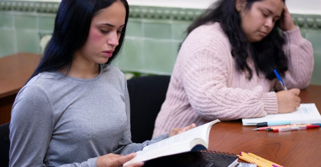 Dos estudiantes inmersos en la lectura y escritura en un aula. Ambiente educativo con libros y material de oficina en la mesa.